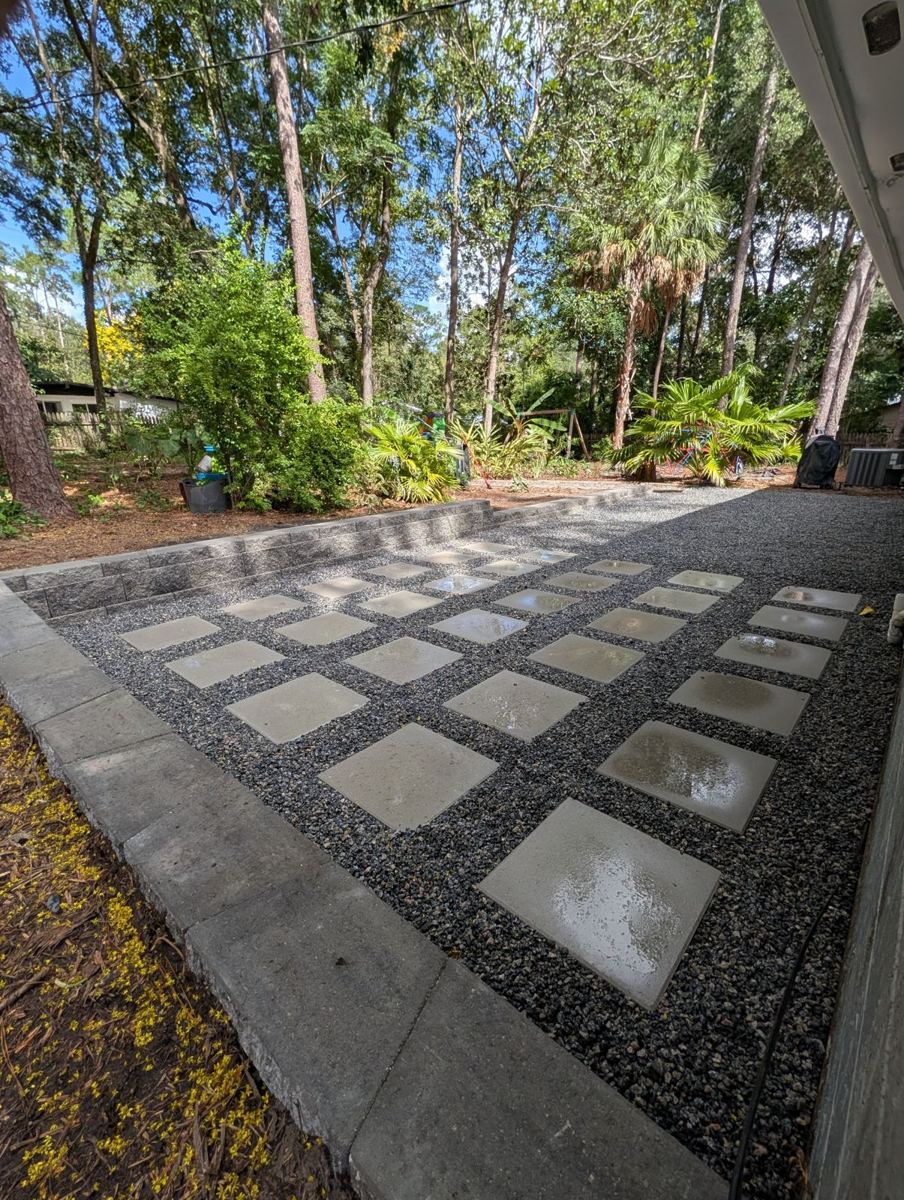 Finished backyard patio with square pavers set in dark gravel and a block retaining wall